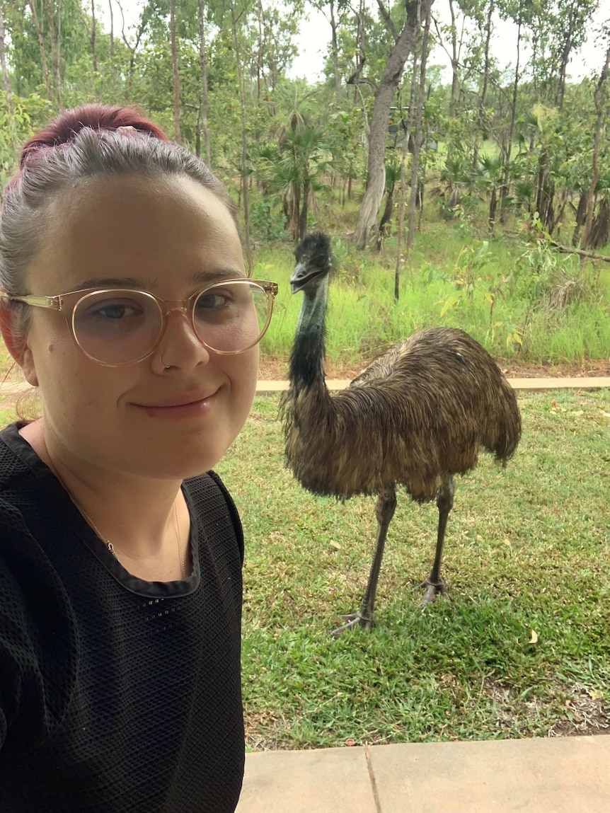 A woman with red hair and glasses smiles at the camera. She is standing outdoors with an emu in the background.