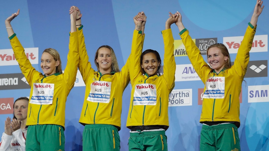 Madison Wilson, Emma McKeon, Kotuku Ngawati and Ariarne Titmus raise each other's hands on the podium.