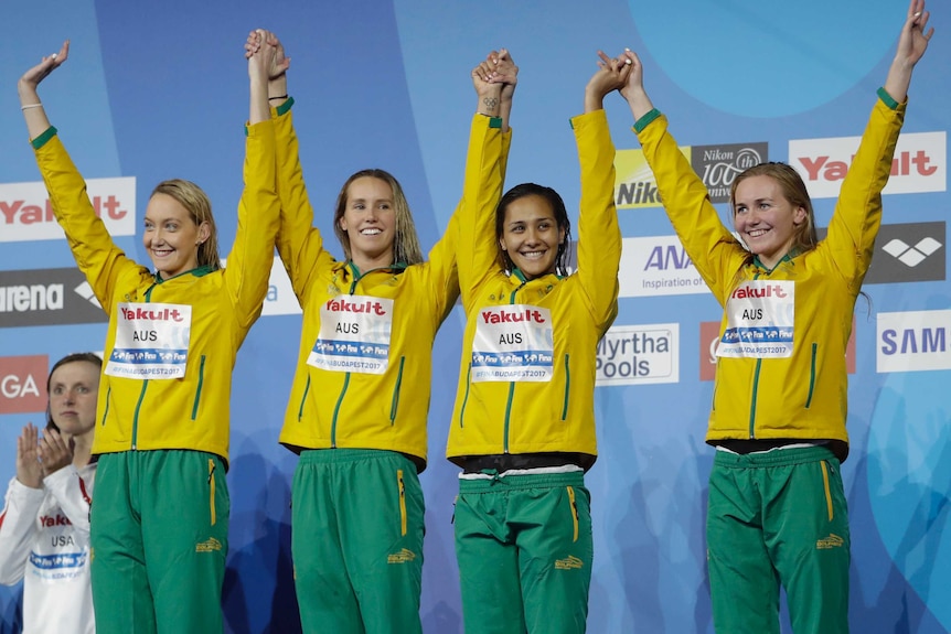 Madison Wilson, Emma McKeon, Kotuku Ngawati and Ariarne Titmus raise each other's hands on the podium.