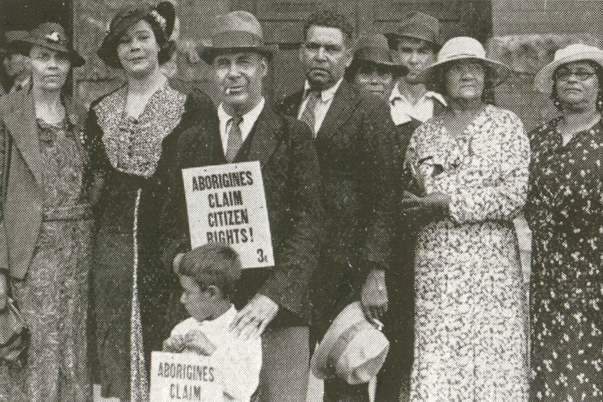 A black and white photo of Aboriginal men and women in 1938, holding a sign that reads 