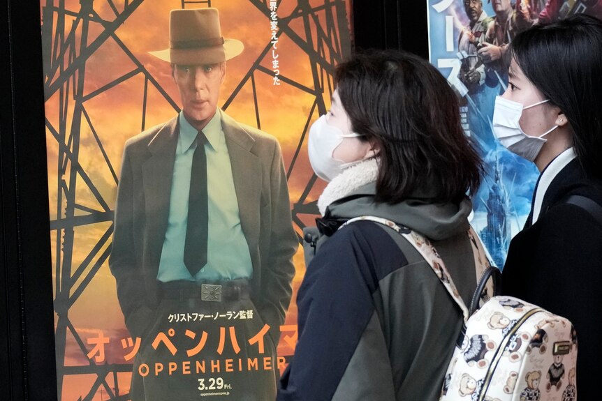 Two young Japanese women wearing face masks looking at a film poster showing a white man in a suit and hat.
