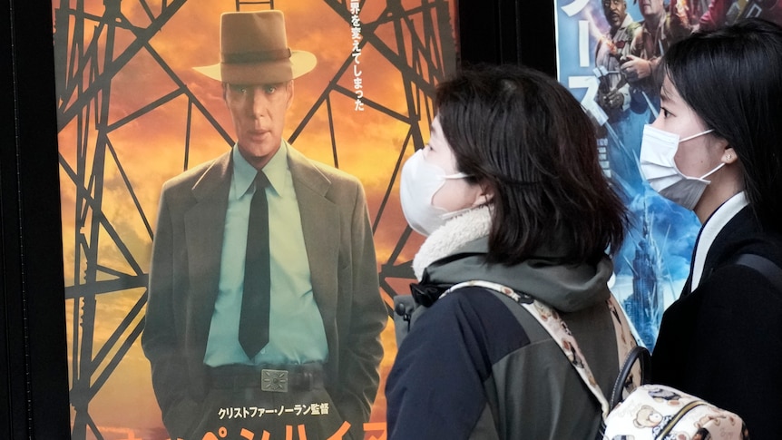 Two young Japanese women wearing face masks looking at a film poster showing a white man in a suit and hat.