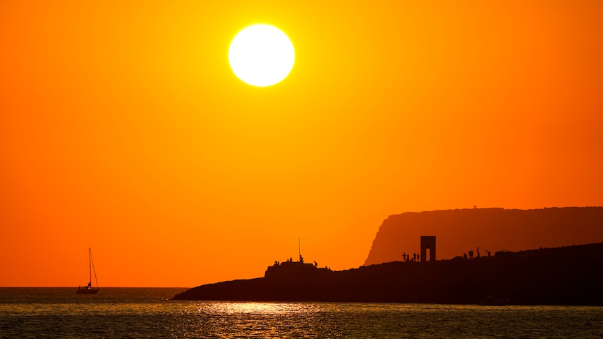 An island and tiny people standing around a statue of a squarish door are silhouetted against a bright orange sky.