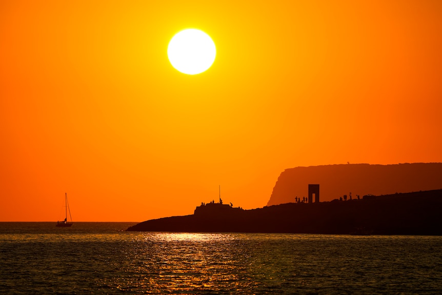 An island and tiny people standing around a statue of a squarish door are silhouetted against a bright orange sky.
