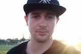 A Selfie-style shot of a young man wearing a flat-brimmed cap standing outside with the sun shining over his shoulder.