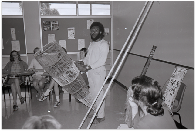 A black and white photo of Eddie Mabo dressed in white showing a class of students how to use a traditional fishing net