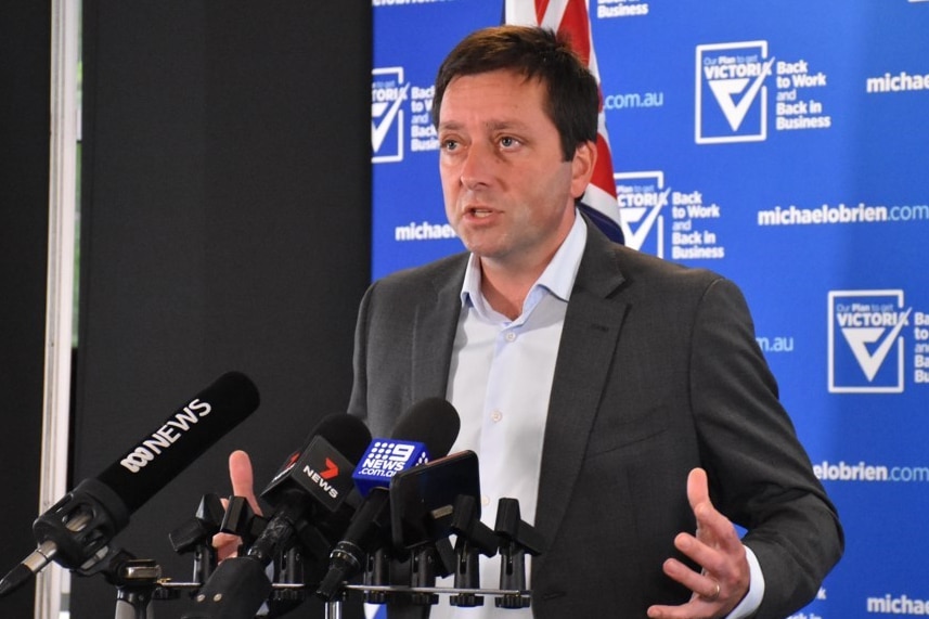 A man in a black jacket and white shirt stands in front of a blue and white background at a press conference.
