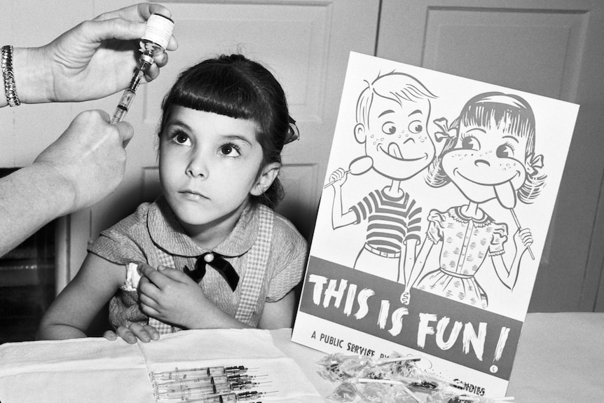 A little girl looks at a needle as she prepares to get the polio vaccine next to a school sign in the 1950s.