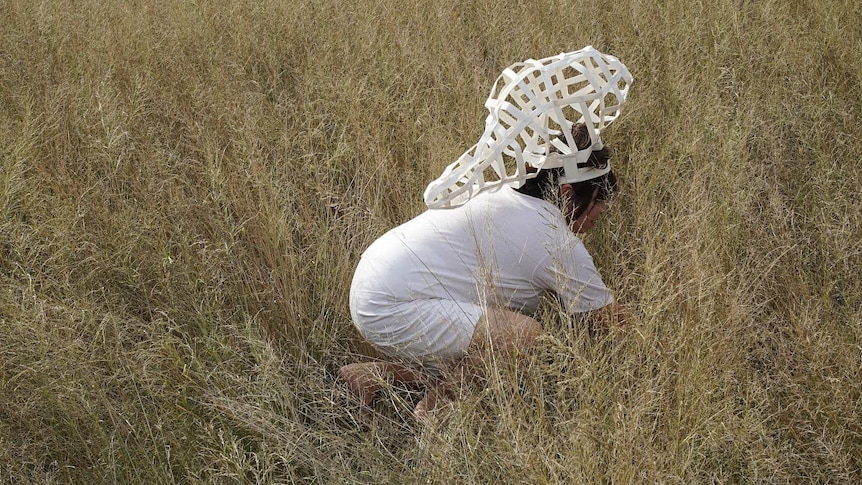 A woman wearing an elaborate hat made of paper crouches in dry grass.