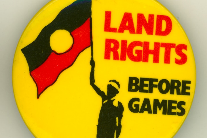 A yellow badge with a figure in silhouette holding the Indigenous flag, with words Land Rights Before Games in red and black.