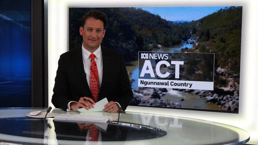 Bourchier at news desk with plasma screen showing ACT ABC News with Ngunnawal Country written underneath.