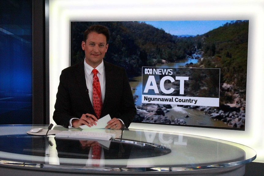 Bourchier at news desk with plasma screen showing ACT ABC News with Ngunnawal Country written underneath.