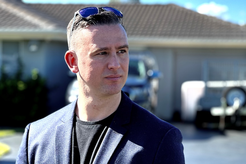 A man in his mid 30s stands in front of a house wearing a blazer and t-shirt with sunglasses on top of his head.