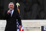 An older man in a coat speaks into a microphone on marble steps in front of a US flag
