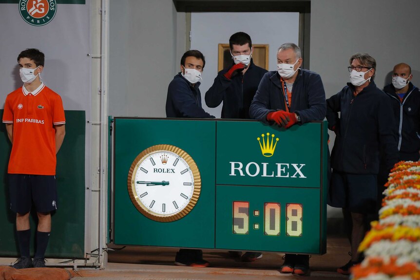 A group of people wearing masks stand behind a French Open clock showing five hours eight minutes.