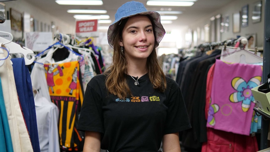 Jess looks at the camera and smiles as she stands among racks of clothes at an op shop. She wears a black t shirt and shorts.