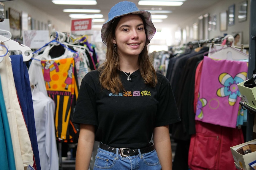 Jess looks at the camera and smiles as she stands among racks of clothes at an op shop. She wears a black t shirt and shorts.