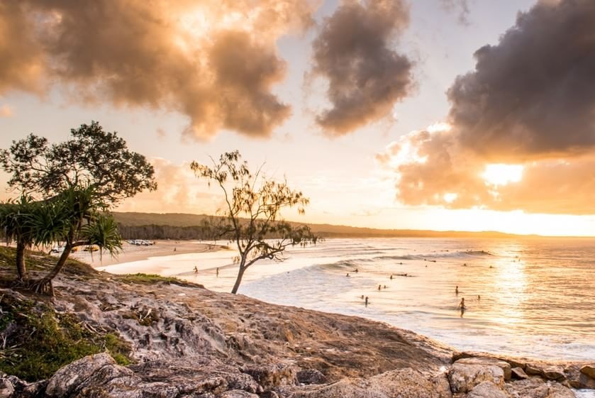 The sunrises over a coastline as people surf.