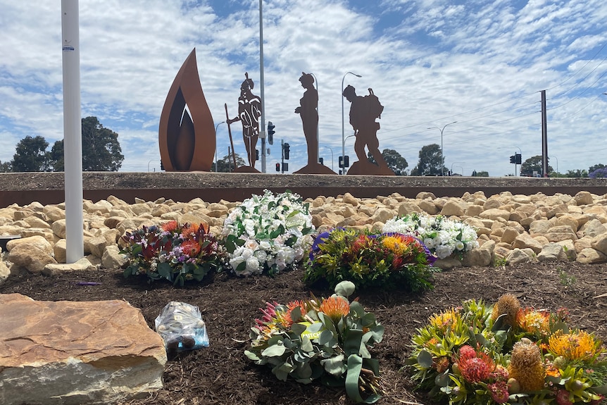 Three figures and a flame shape made out of metal with rocks and native flowers in front of it