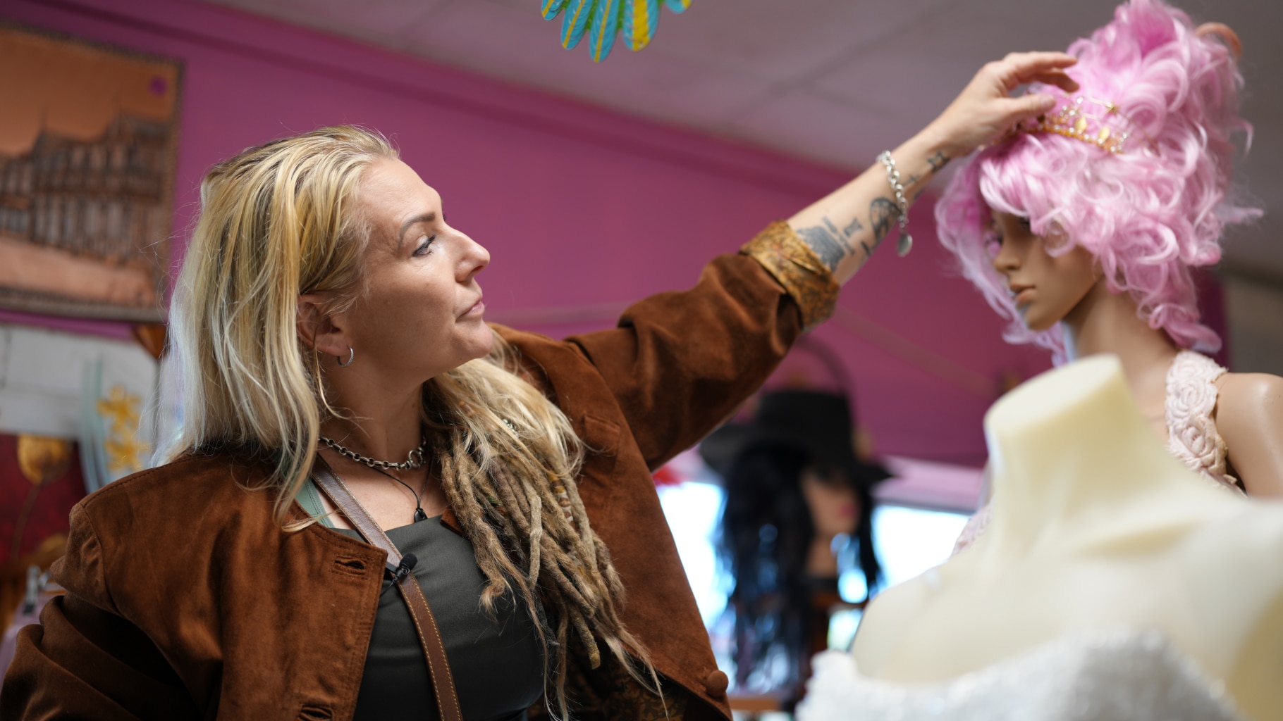 A blonde woman with dreadlocks reaching out her left hand to adjust a headband on a mannequins head.