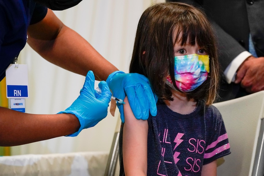 A young girl wearing a tie-dye rainbow face mask sits still while a nurse delivers a vaccine by injection