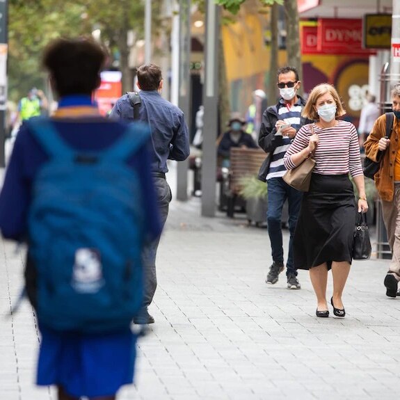 A blurred school child carries their bag while a trio of adults wearing face masks walk by on an inner city street.