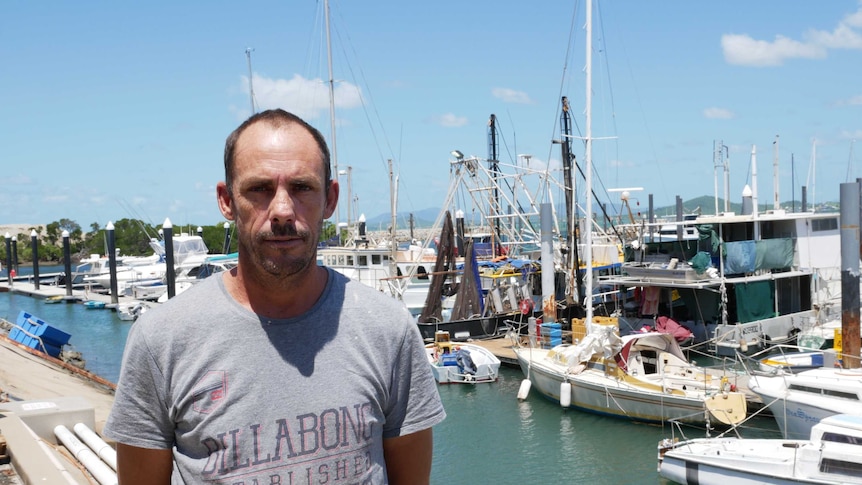 A middle-aged man with a receding hairline stands on a wharf in front of a motley regatta of moored fishing vessels.