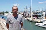 A middle-aged man with a receding hairline stands on a wharf in front of a motley regatta of moored fishing vessels.