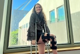 A woman stands on crutches with her right knee held up smiling at the camera. The knee is in a blue knee brace
