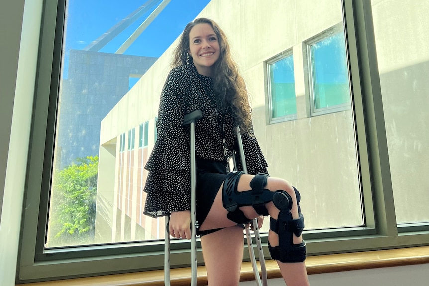 A woman stands on crutches with her right knee held up smiling at the camera. The knee is in a blue knee brace