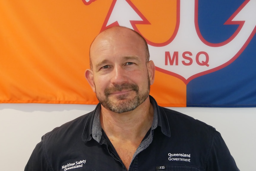 A man with stubble looks at the camera. There is an orange and blue maritime flag in the background.