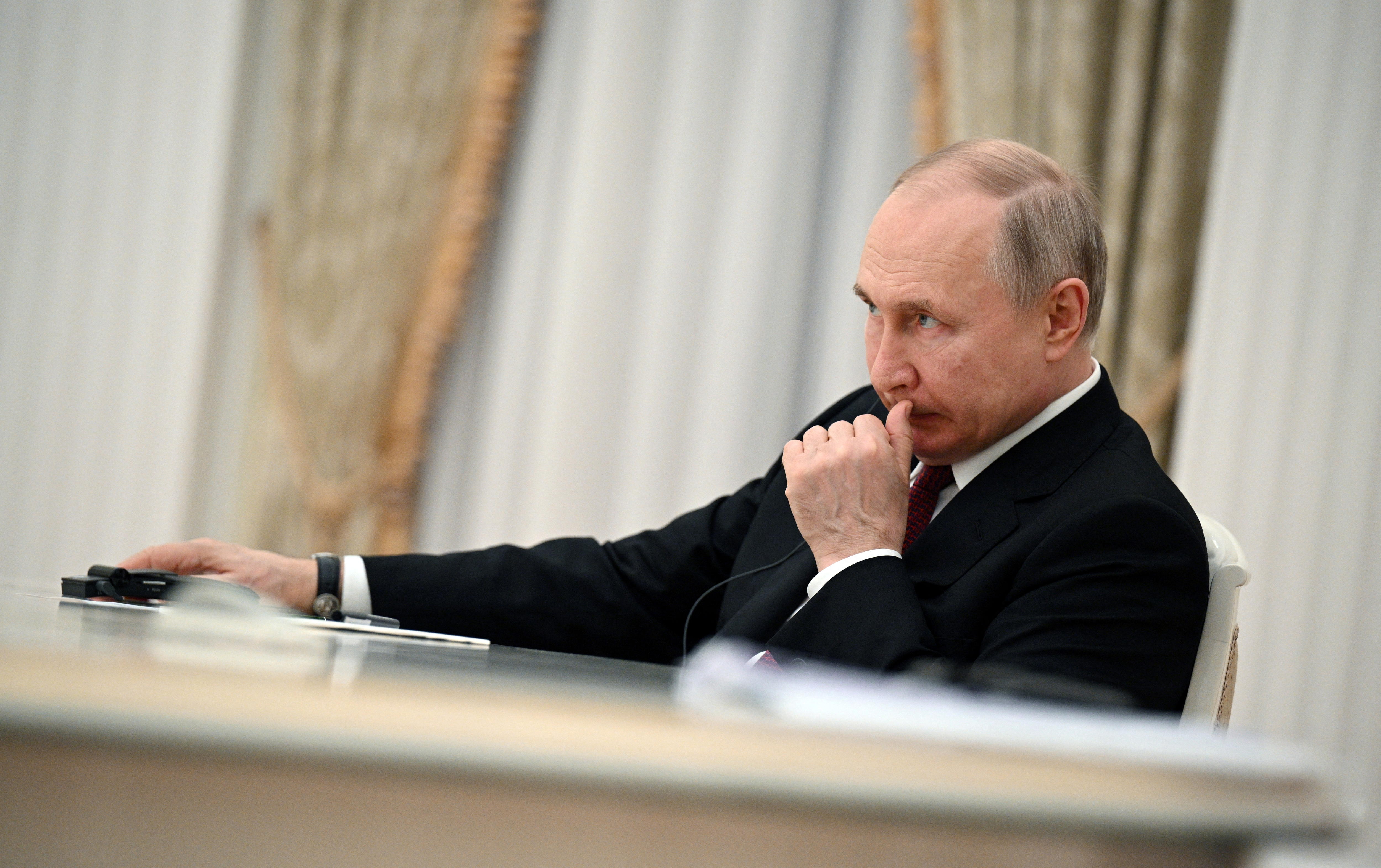 Vladimir Putin rests one arm on a desk in front of him, scratching his upper lip with a concerned expression on his face