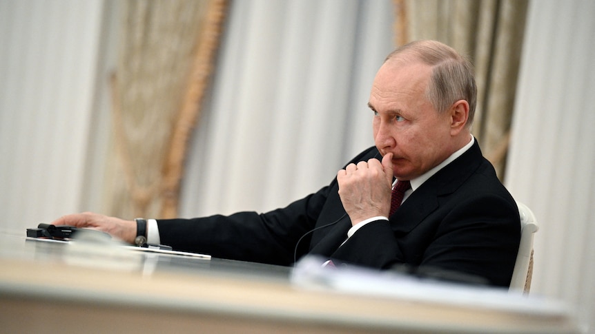 Vladimir Putin rests one arm on a desk in front of him, scratching his upper lip with a concerned expression on his face