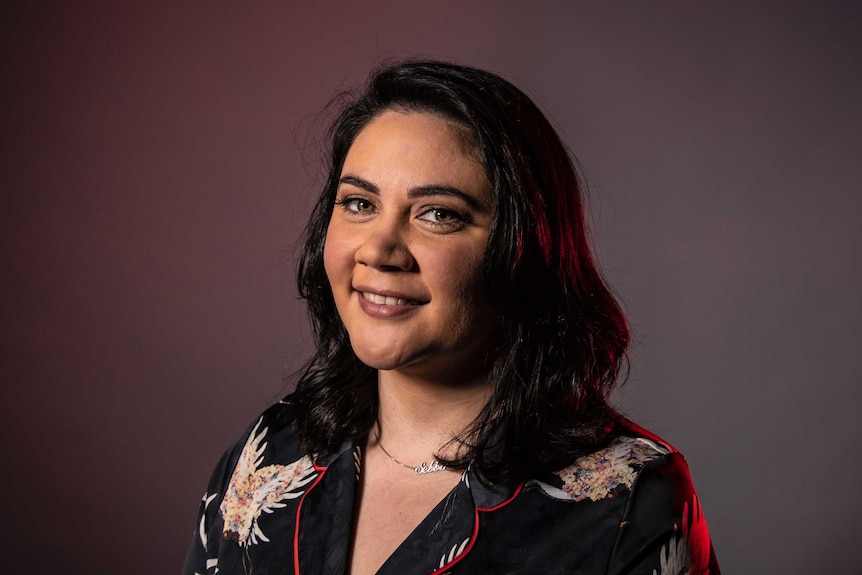 A woman with dark hair and green eyes wears black patterned shirt and poses in front of grey with light magenta on left side.