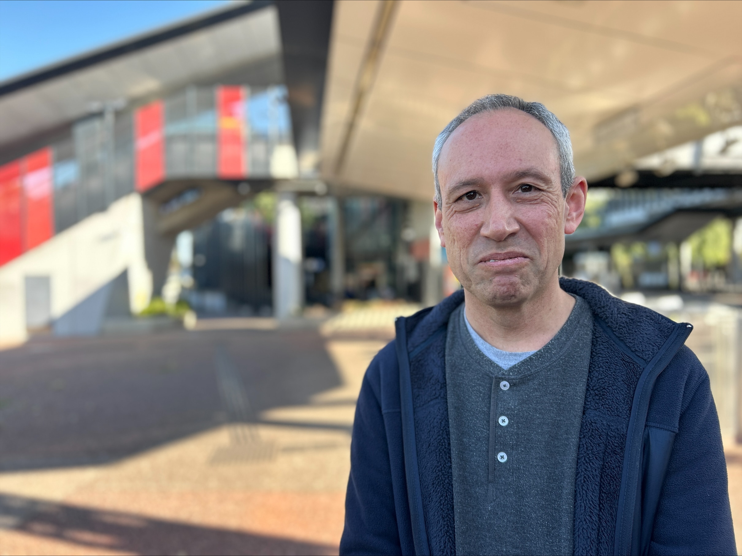 Un hombre con cabello gris, camisa gris y suéter oscuro mira fijamente a la cámara con una sonrisa en los labios cerrados.