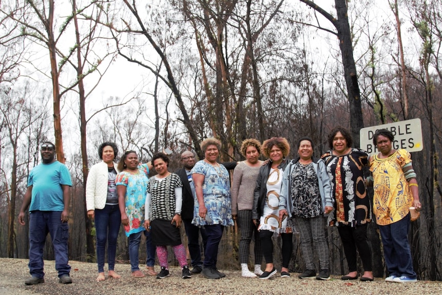 A group of 9 women and 2 men from PNG community stand on a road in front of charred trees.