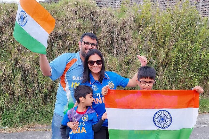 An Indian family, wearing blue, smile and wave Indian flags on a quiet road with greenery behind them