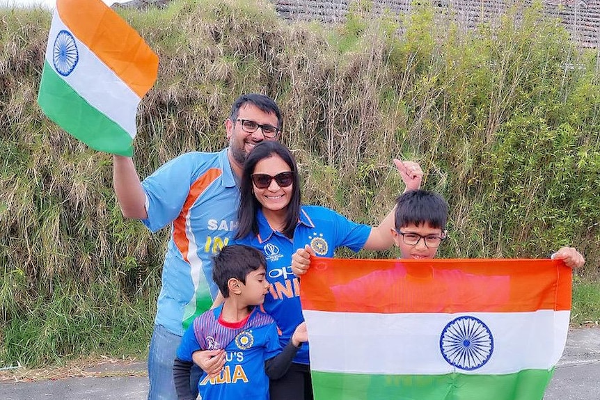 An Indian family, wearing blue, smile and wave Indian flags on a quiet road with greenery behind them
