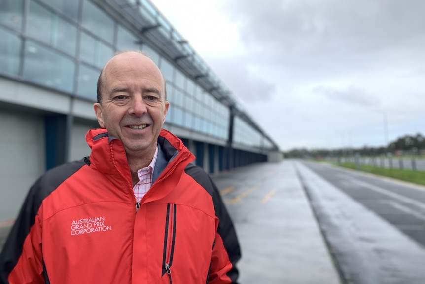 Andrew Westacott smiles, dressed in a red rain jacket out next to a motor racing track.