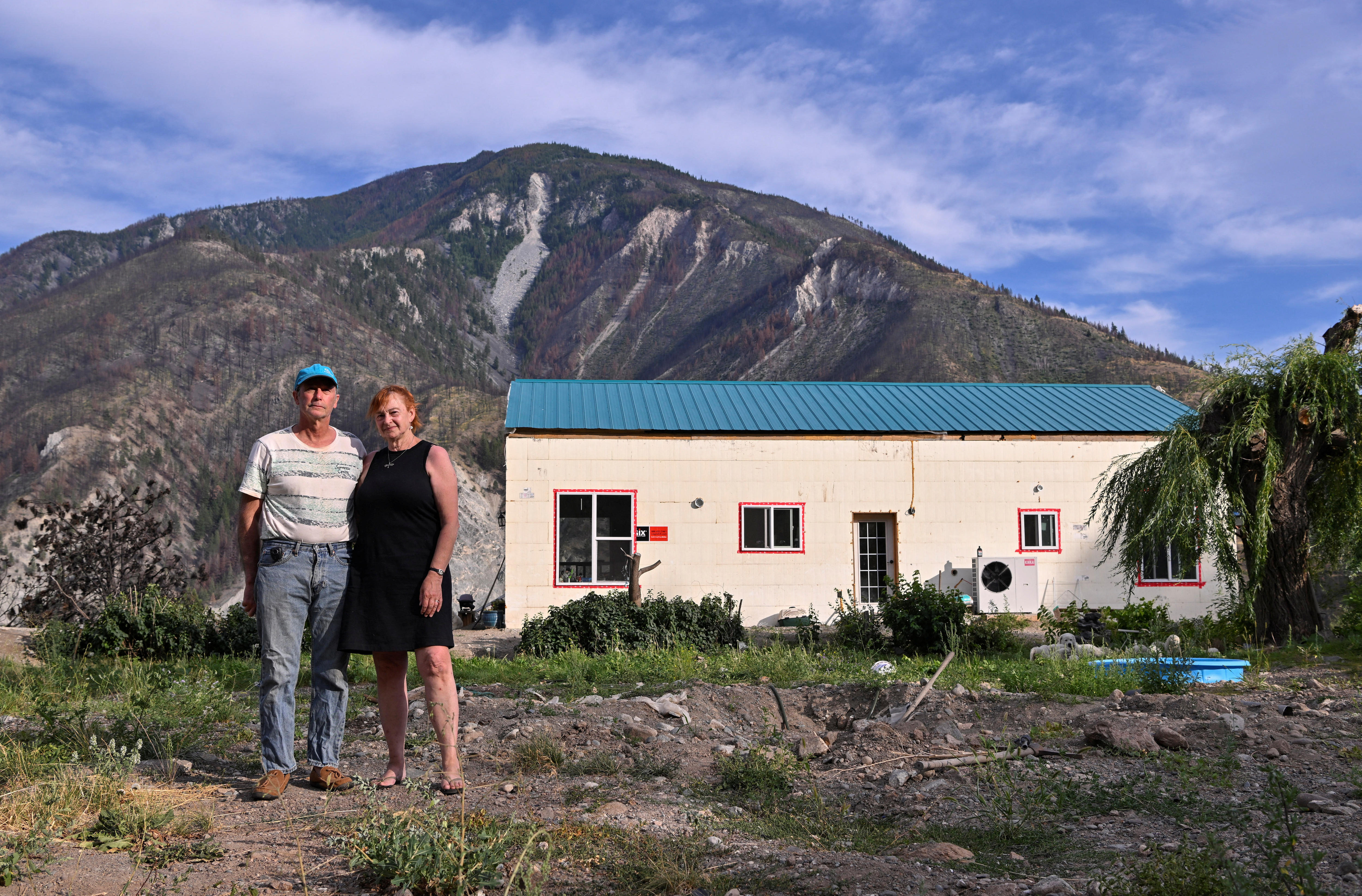 A middle-aged couple stand in a patchy front yard in front of a basic white brick building with a blue roof.