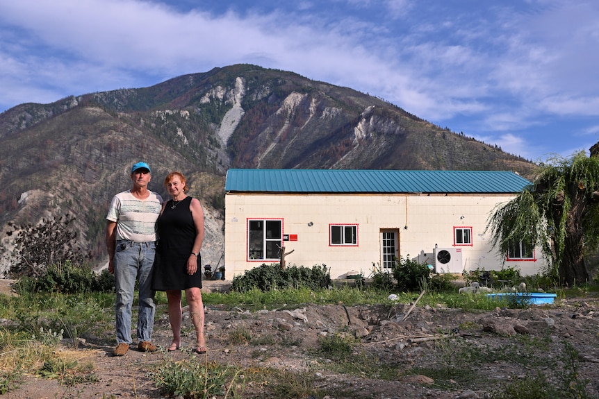 A middle-aged couple stand in a patchy front yard in front of a basic white brick building with a blue roof.