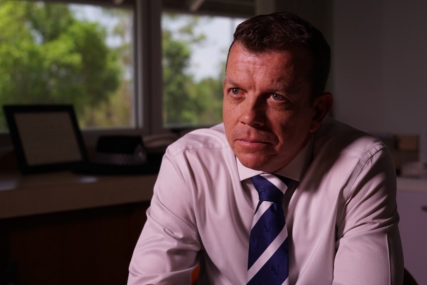 A middle-aged man in a business shirt and tie sits at a table leaning on his elbows.