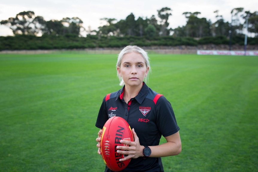Courtney Ugle stands on a green football field, holding a footy and looking into the lens.