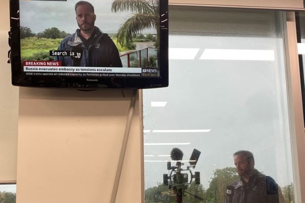 Man standing outside a window of a room with a camera doing a cross with a TV screen inside room showing him on air.