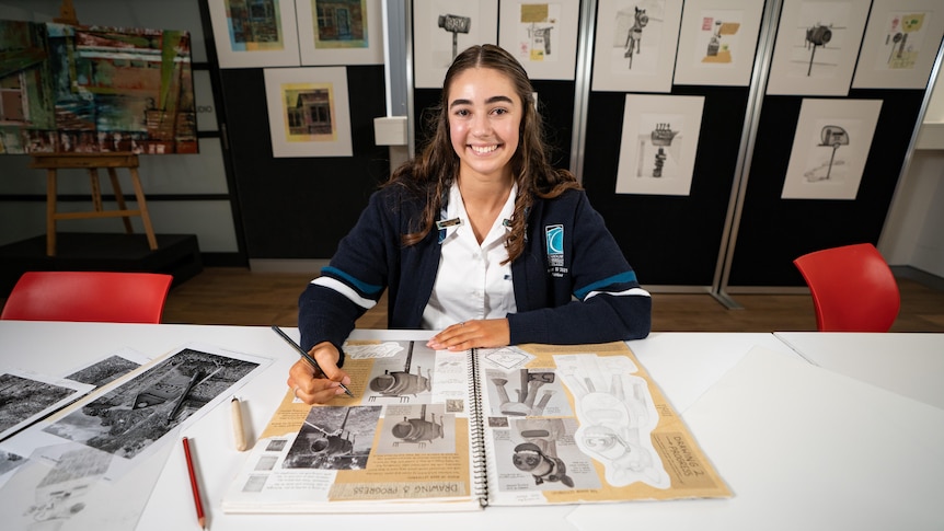 Amber smiles brightly at the camera while writing in a sketchbook, different drawings are hanging on the wall behind her.