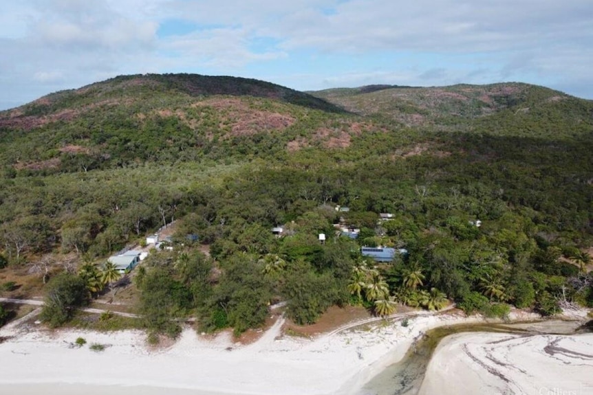 Some small buildings photographed from the air hidden by rainforest backing onto a white sandy beach 