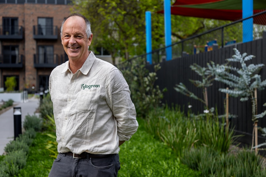 A man wearing a beige buttoned shirt standing surrounded by a green garden.