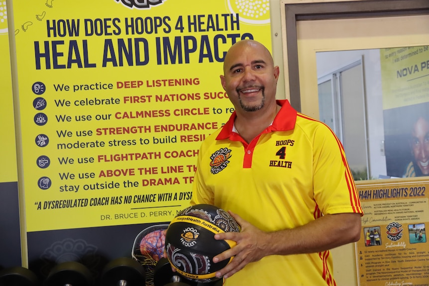 A man holding a basketball in front of a sign with Hoops 4 Health printed on it.
