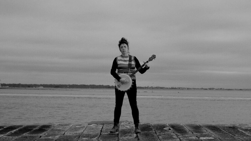 A landscape shot of a woman standing on a dock holding a banjo and looking very serious.