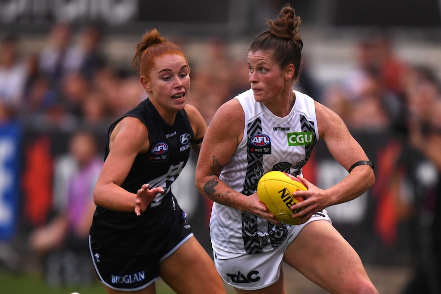 A Collingwood AFLW player runs with the ball in two hands in front of a Carlton opponent.